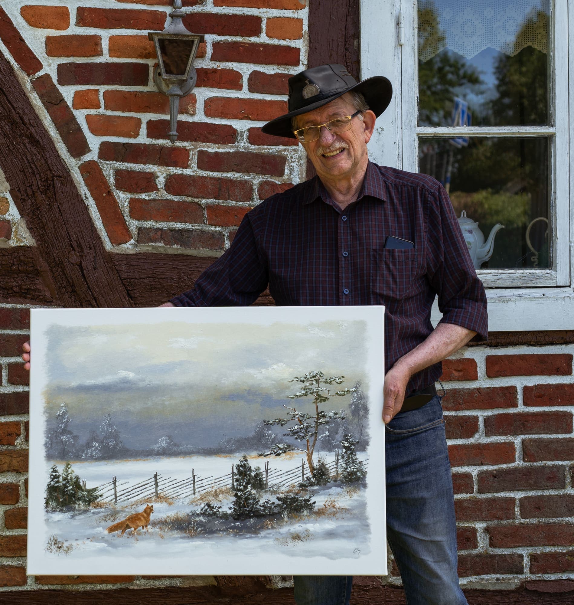 Konstnären Ingvar Stendahl står framför sitt korsvirkeshus på Skomakaregården och håller upp en av sina målningar.